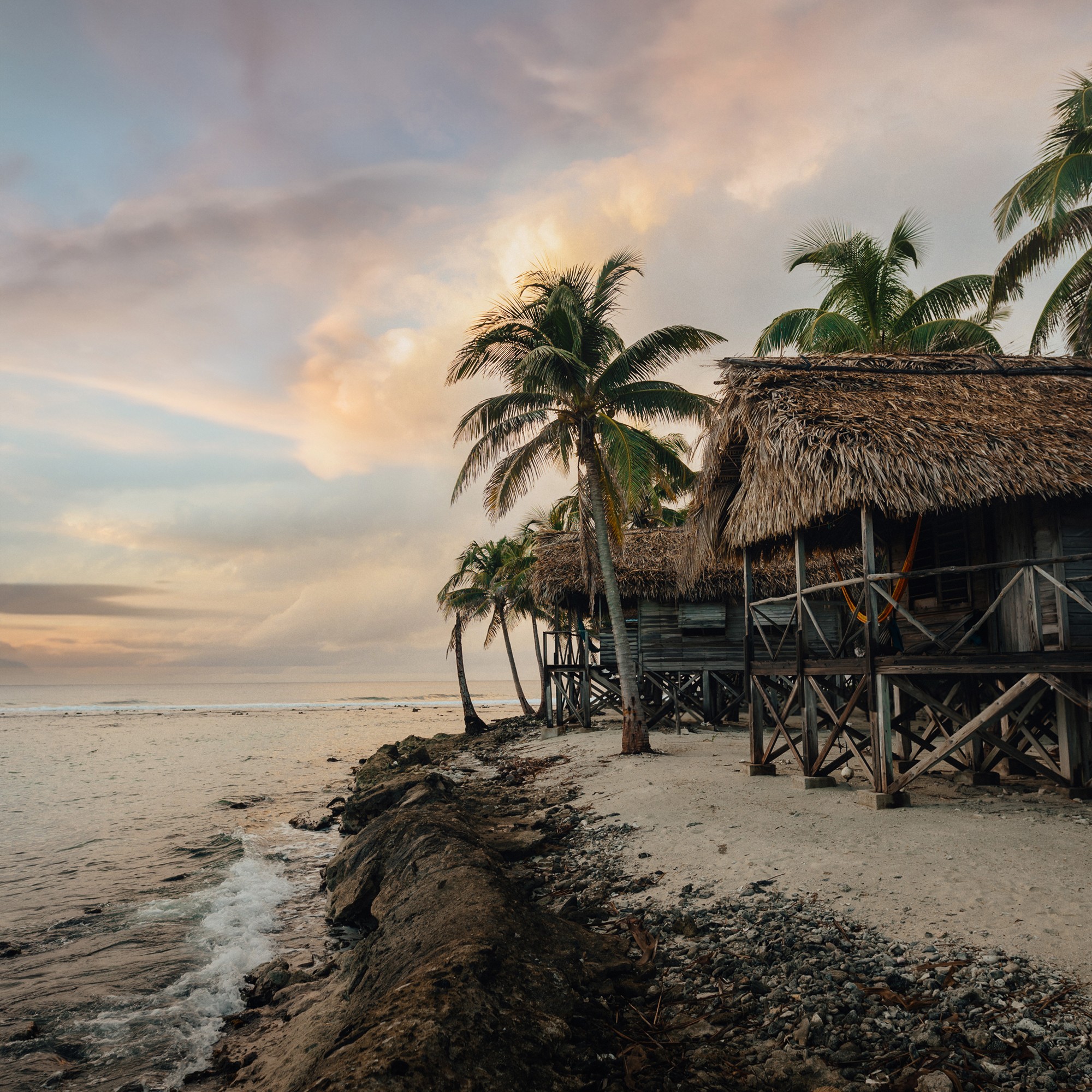 Rustic overwater beach huts with palm trees on a tropical coastline in Belize at sunrise.