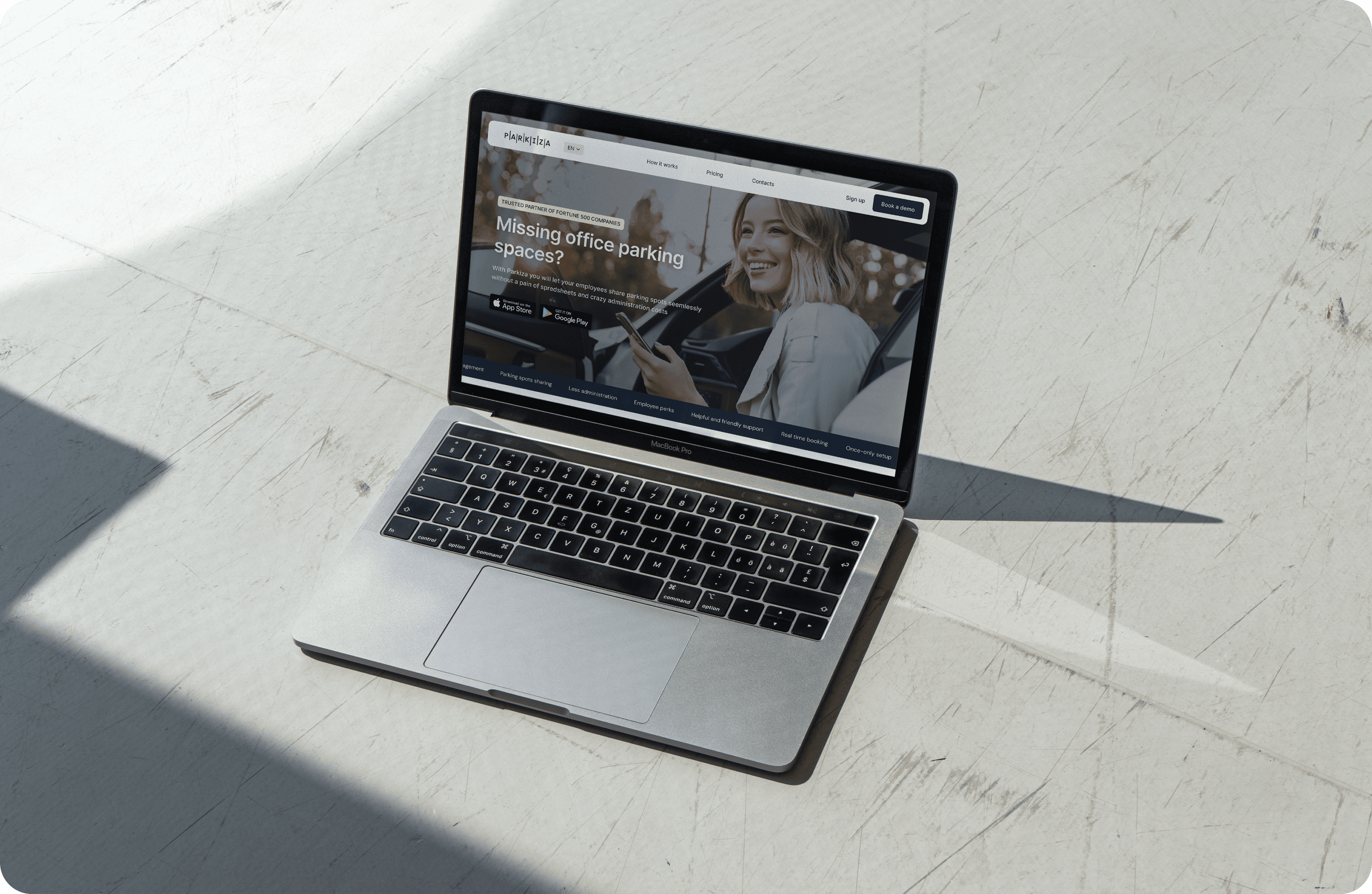 A MacBook Pro laptop on a white marble surface displaying the Parkiza website. The website shows a banner with a smiling woman using her phone and text that reads: 'Missing office parking spaces? Parkiza helps you and your employees share, track and manage parking spaces seamlessly from your phone or desktop.' There are buttons for downloading the app from the App Store and Google Play. The top navigation bar includes options for 'How it works,' 'Pricing,' 'Resources,' 'About,' 'Contacts,' and a 'Sign up' button. There is also a 'Book a demo' button on the right side of the banner.