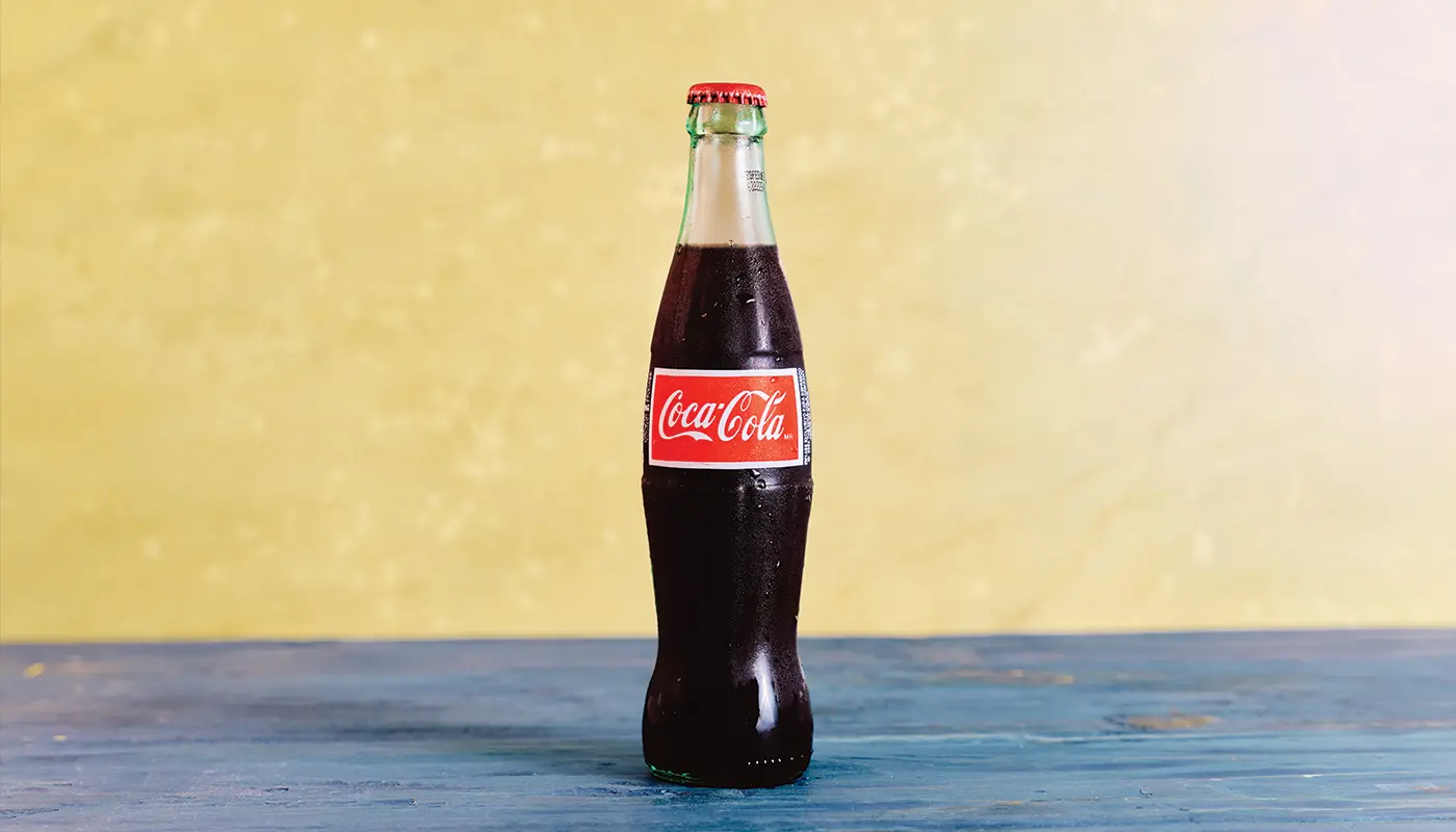 A glass Coca-Cola bottle on blue wooden boards against a yellow wall.