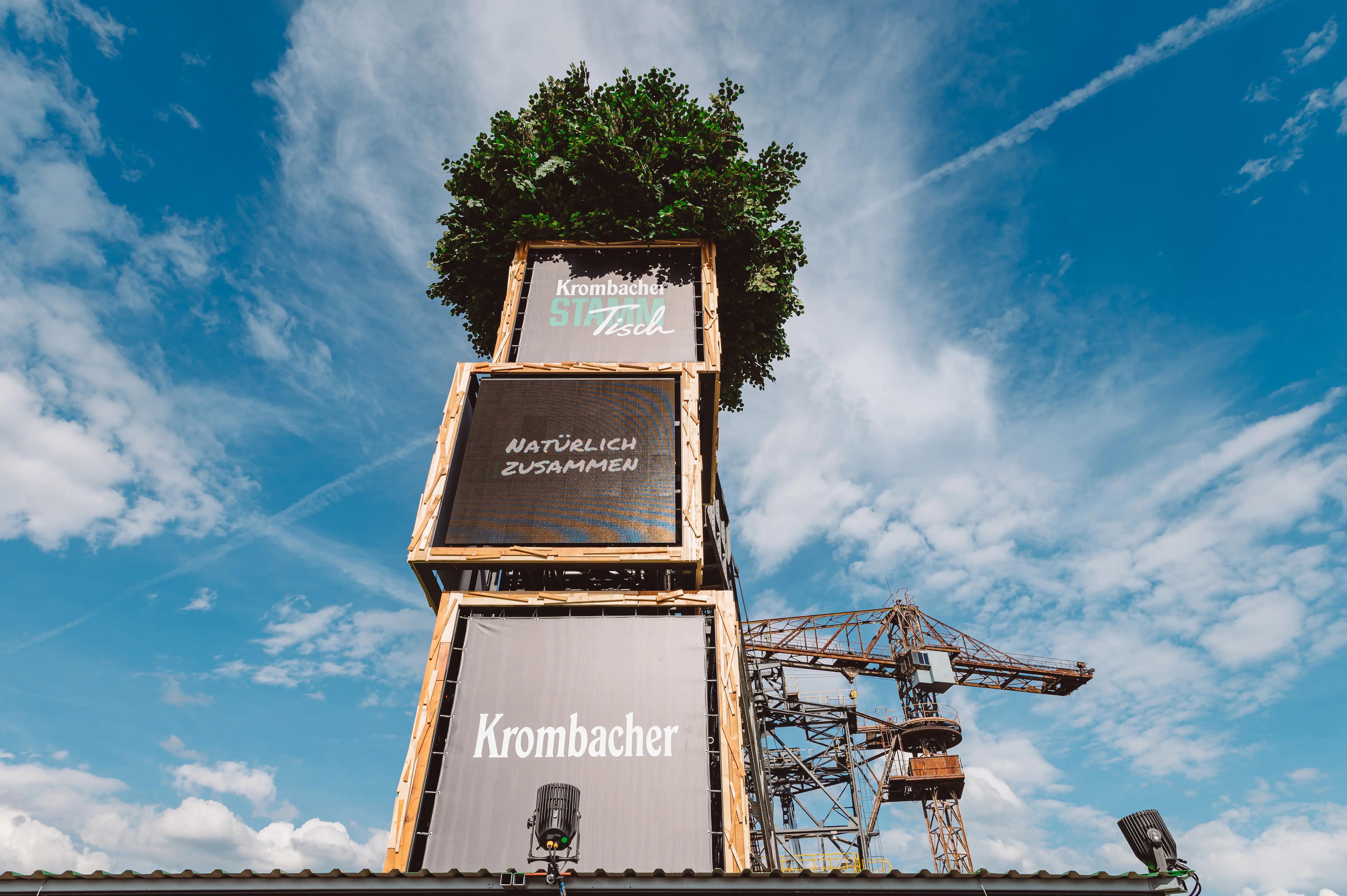 Low-angle view of the KROMBACHER tower with Natürlich Zusammen lettering and a tree on top, against a blue sky with the Ferropolis crane.