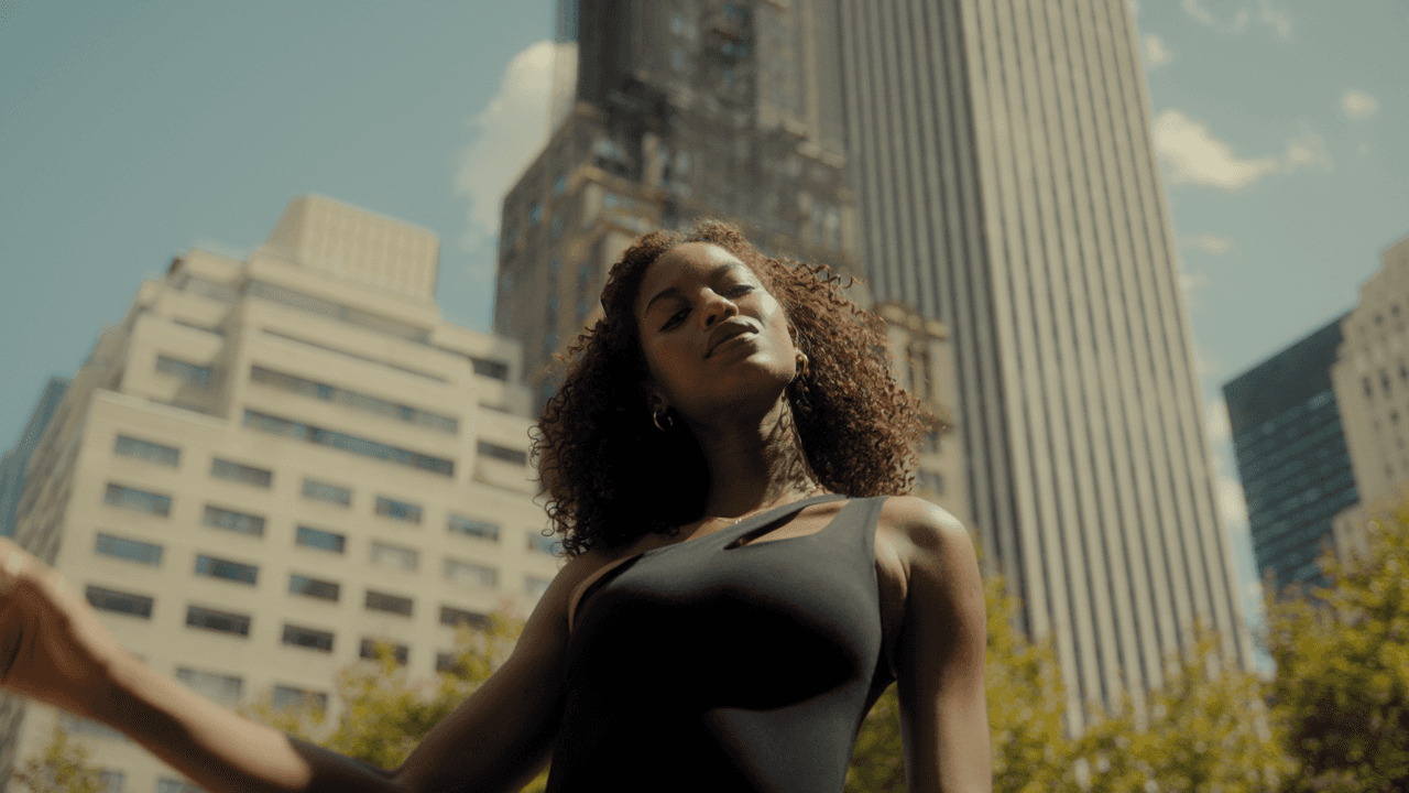 A model standing in front of tall city skyscrapers, basking in sunlight, with her head tilted upward