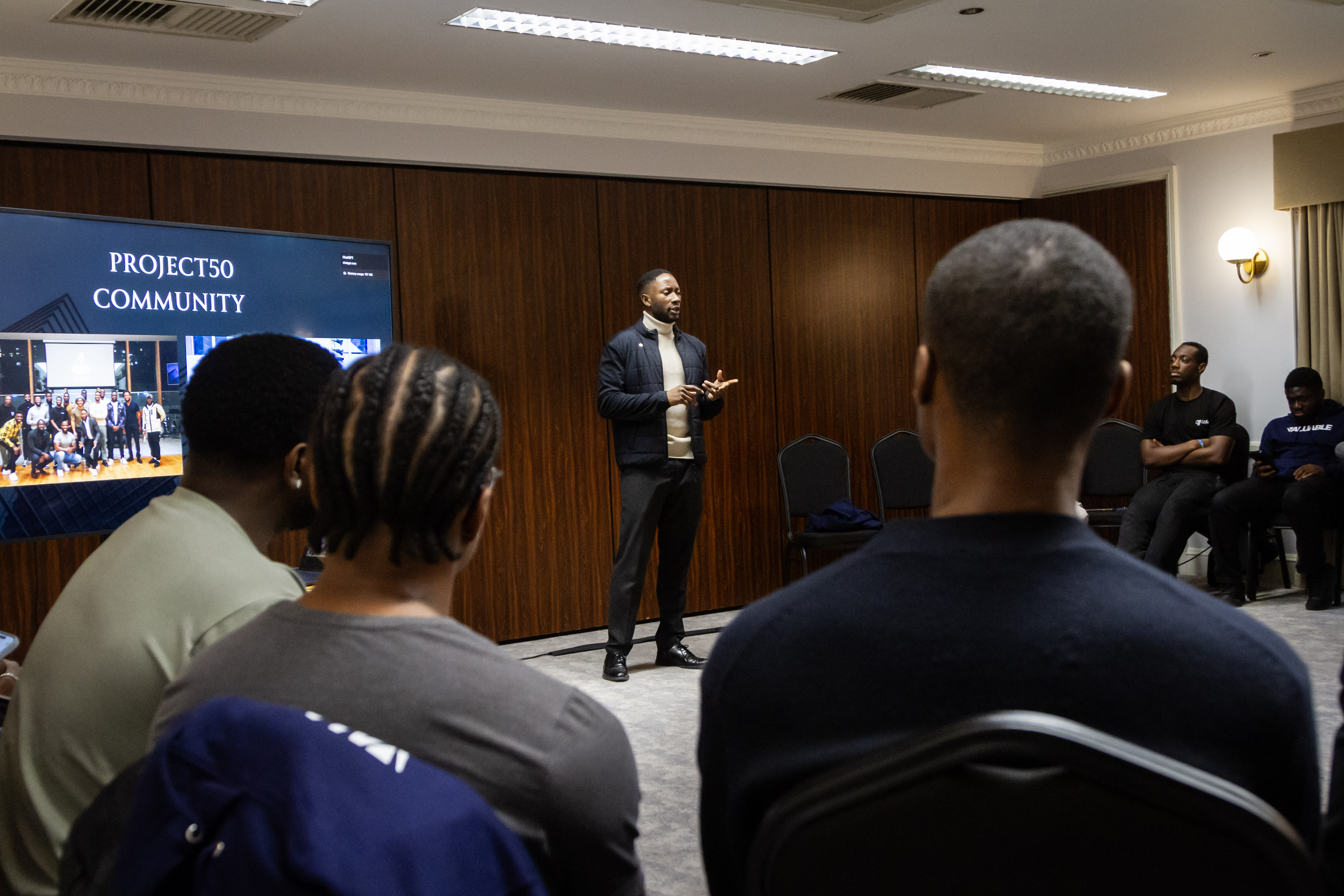 Man standing and giving a talk in a conference room with men gathered sitting down on chairs arranged in a circle.
