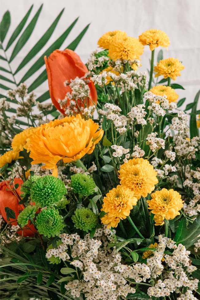 Detall d’un ram variat amb crisantems grocs, ranuncles taronja, tulipes, flor d’arròs blanca i craspèdia verda, en una composició exuberant i vibrant.