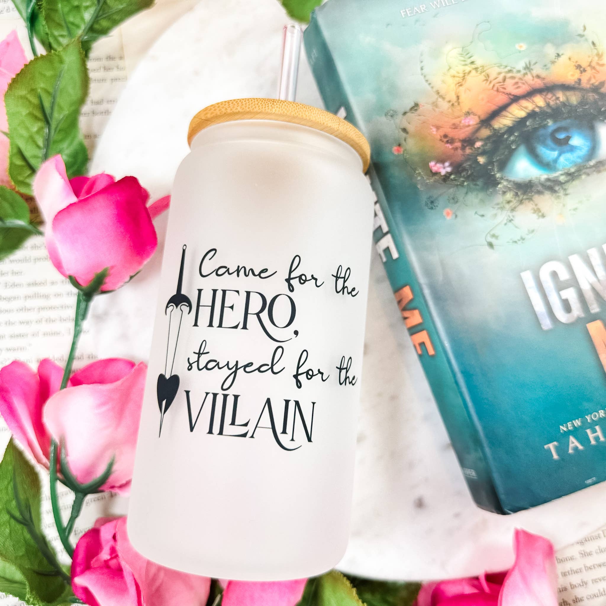Frosted glass cup reading 'Came for the hero, stayed for the villain' surrounded by pink roses and book pages