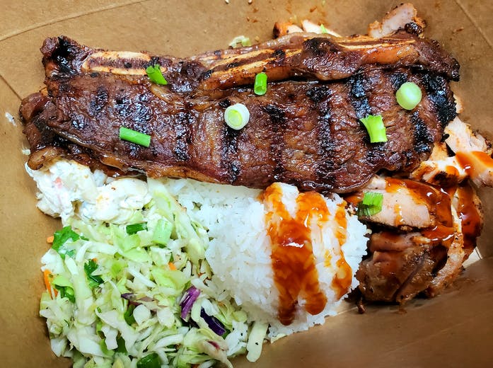 A takeout container filled with two large grilled Angus short ribs, Huli Huli chicken, two scoops of Jasmine rice, Hawaiian mac salad, and Asian slaw.
