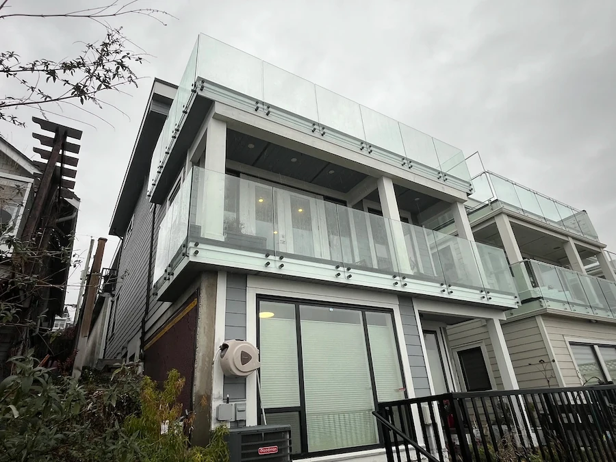 Modern two-storey home with frameless glass balcony railing and exposed timber posts, installed on a sloped property in British Columbia.