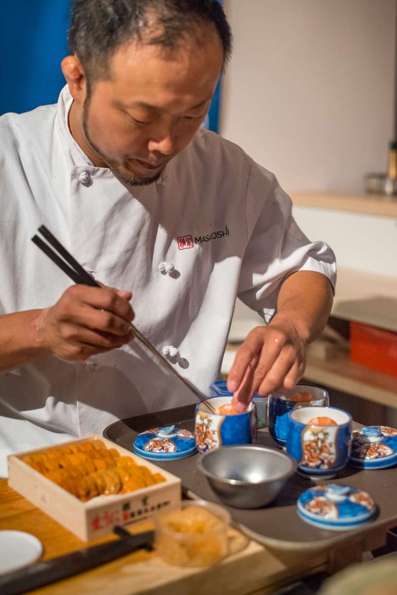 Asian chef carefully arranges colorful, sweet pastries on a wooden tray under bright indoor lighting.