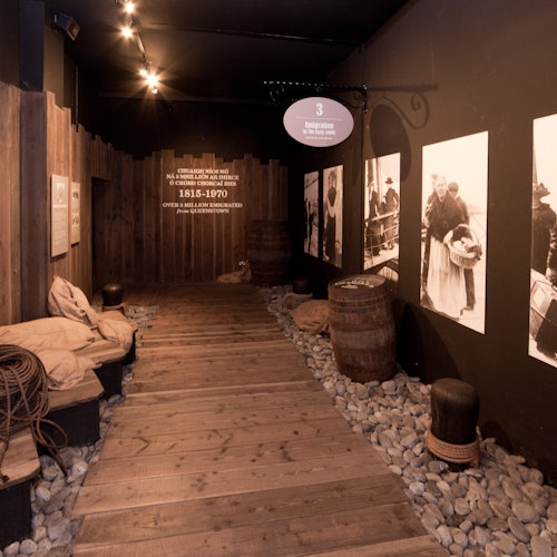 Un chemin en bois entouré de tonneaux, de sacs et de photographies historiques affichées sur les murs dans un espace d'exposition faiblement éclairé.