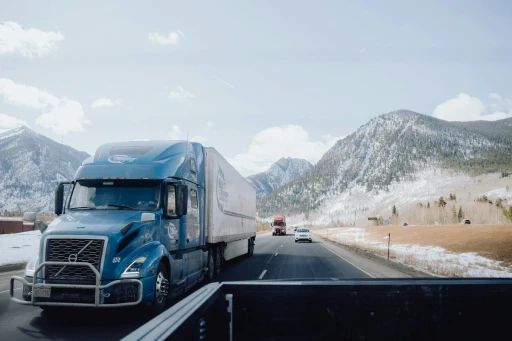Semi-truck driving on a highway through mountainous terrain during winter conditions.