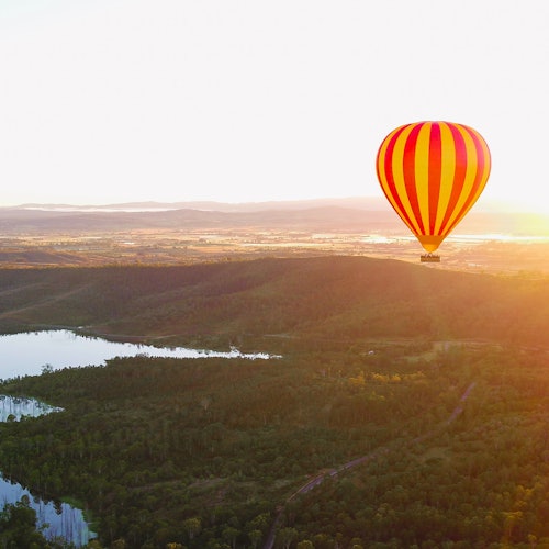 Een rood-gele heteluchtballon zweeft over een landschap met groene heuvels, bomen en water bij zonsondergang.