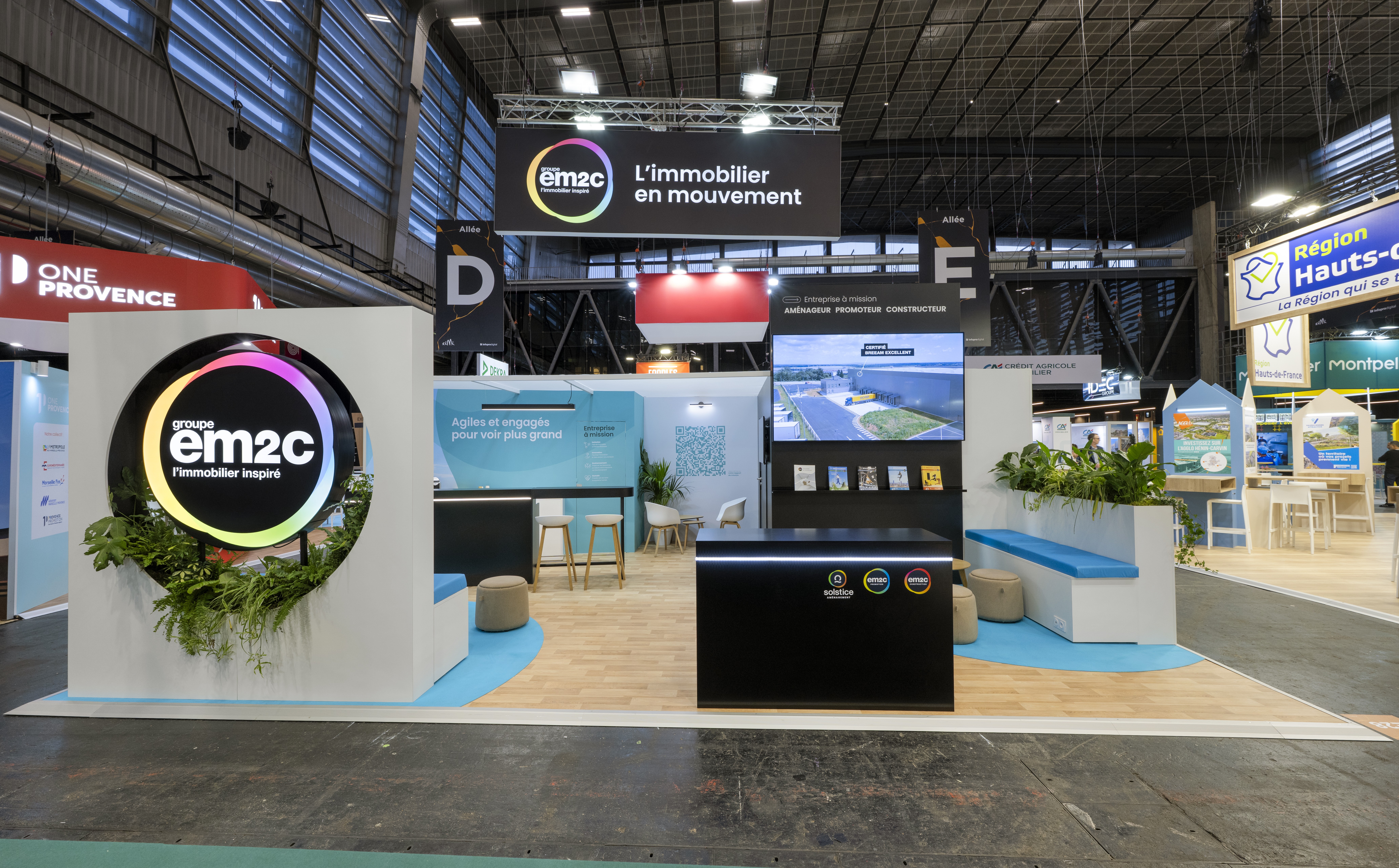 Stand EM2C SIMI 2025 Paris conçu par Expace – vue d'ensemble du stand avec médaillon logo lumineux, structure bicolore noire et bleue et espace conseil immobilier professionnel