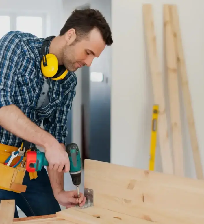 Carpenter using a cordless drill to attach a metal bracket to a wooden plank.