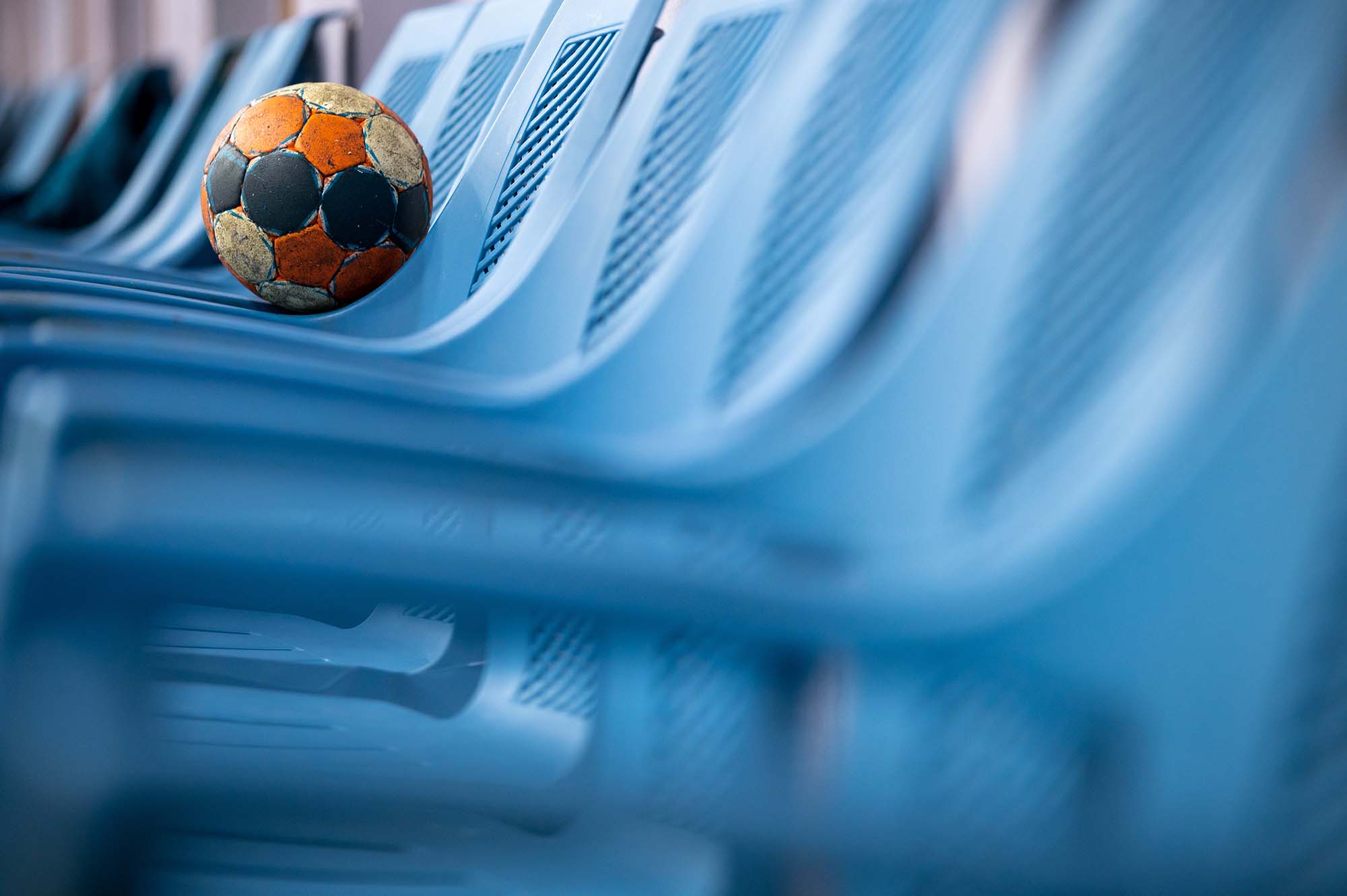 A worn orange and black handball resting on an empty blue stadium seat among a row of vacant chairs.