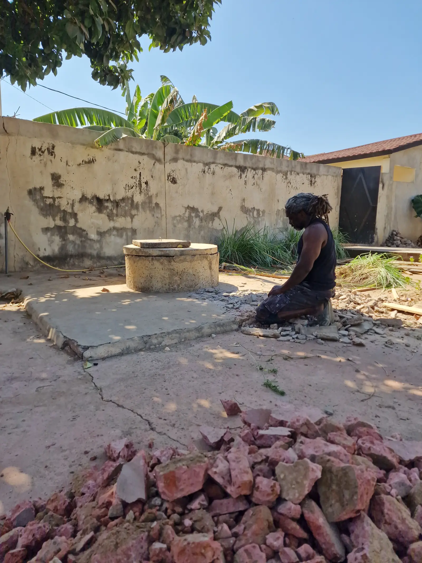 A person kneels on the ground near a stone structure, surrounded by a sunny outdoor setting with trees.