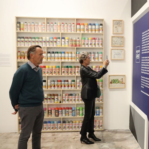 画廊中两个人；一位女士正在拍摄墙上的展品，另一位男士则站在摆满喷漆罐的架子旁。