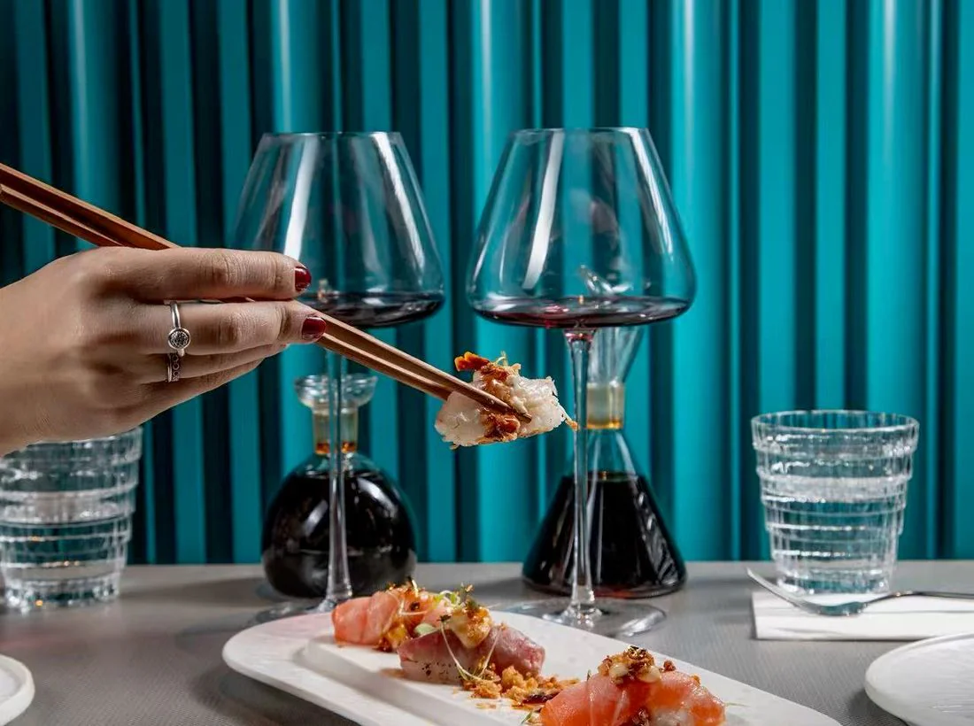 Un sashimi de saumon pris à la baguette, devant deux verres de vin et le rideau de velours