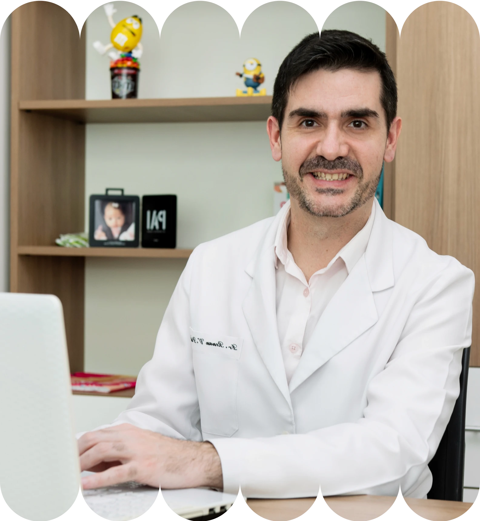 O Dr. Renan, médico de barba rala, usando jaleco branco sobre camisa rosa, sentado em uma mesa de escritório e digitando em um laptop. Prateleiras com decorações infantis, como Minions e um porta-retratos de "Pai", estão ao fundo.