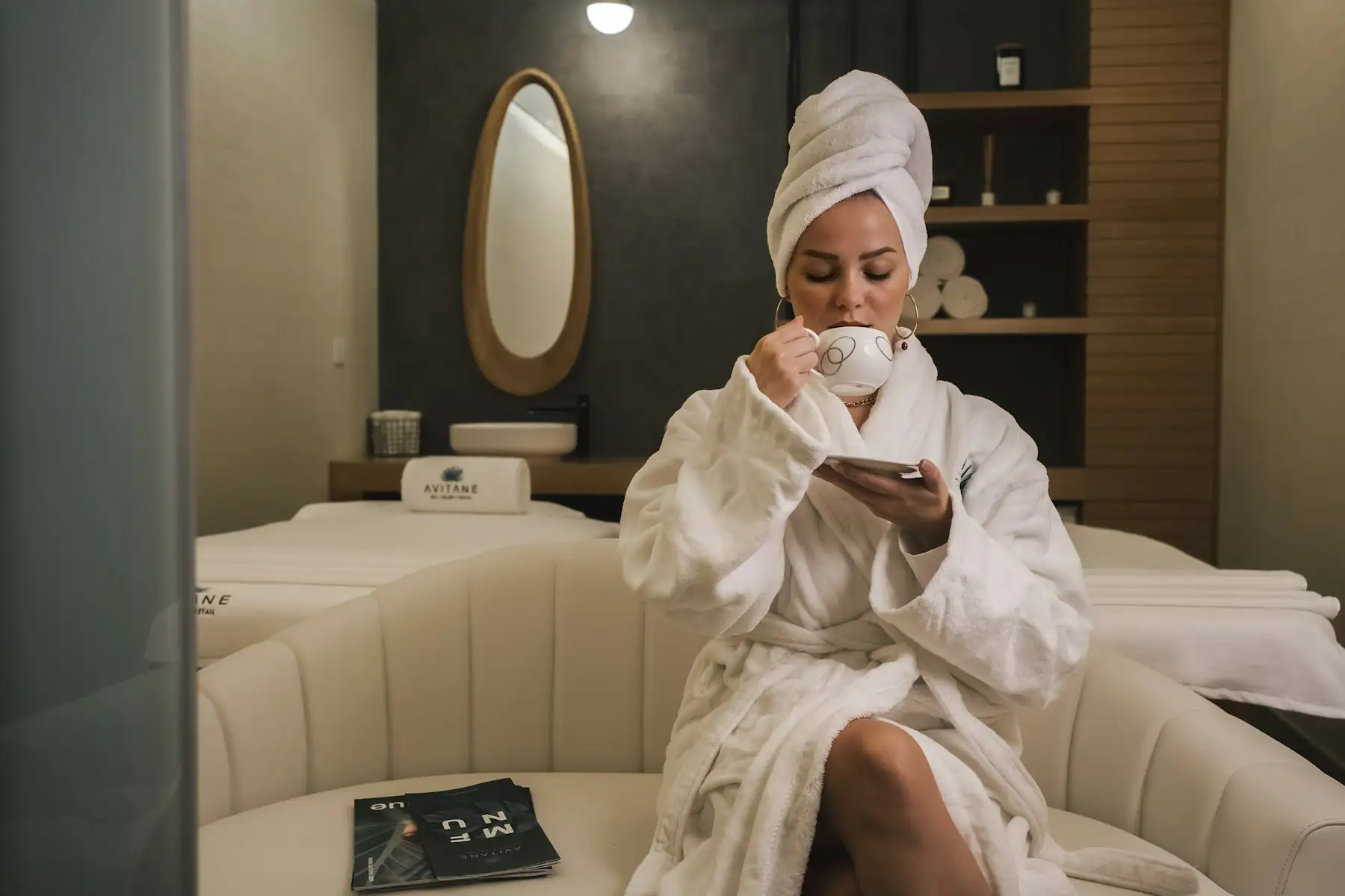 A woman in a white robe and hair towel sitting on a couch with a cup