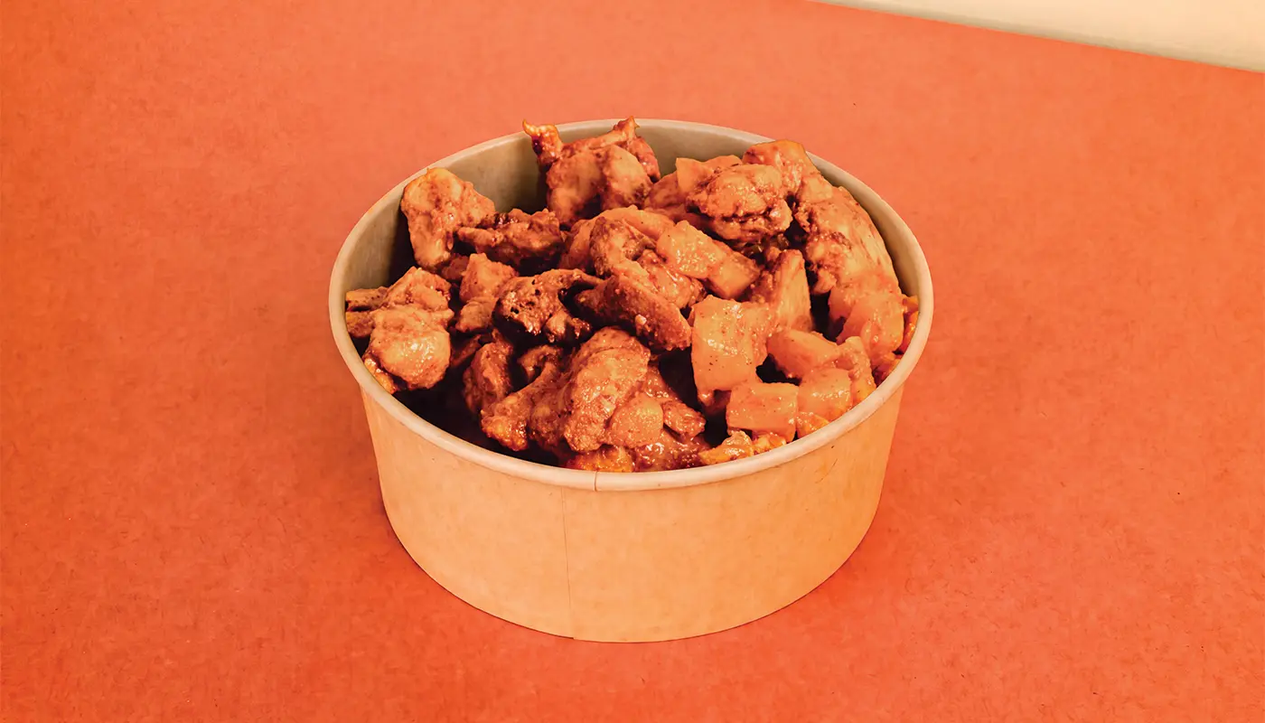 A kraft-paper bowl filled with reddish-orange grilled chicken pieces, charred edges, and green herb garnish.
