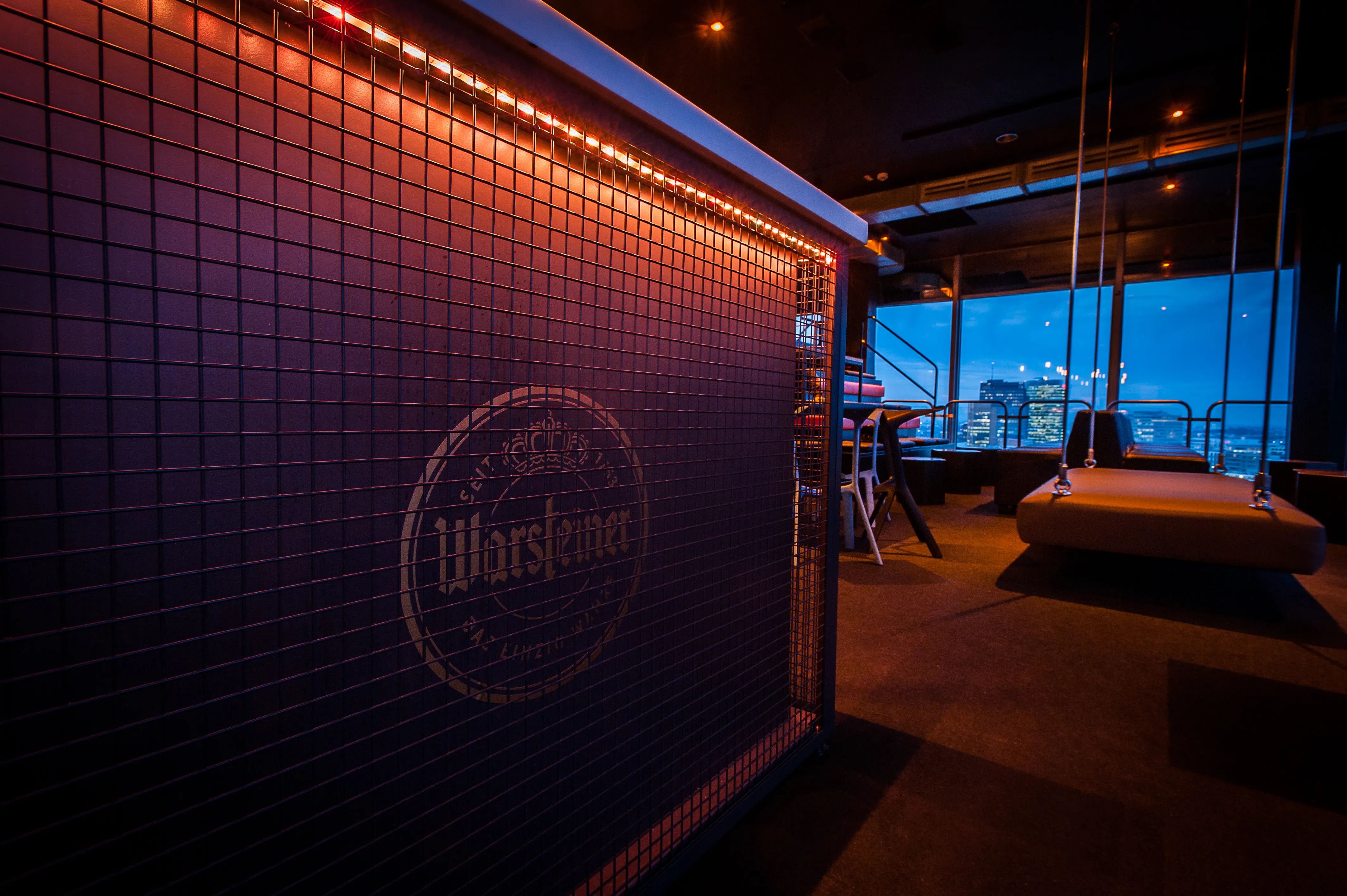 Red-lit metal mesh partition with engraved Warsteiner logo at SOLAR SKY LOUNGE, next to a hanging swing-style daybed in front of night-time skyline windows.