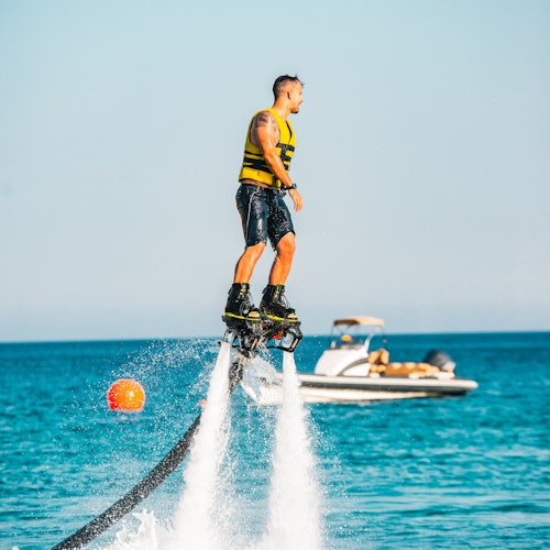 A man in a yellow life jacket is flyboarding over water, with a boat and a red buoy in the background.