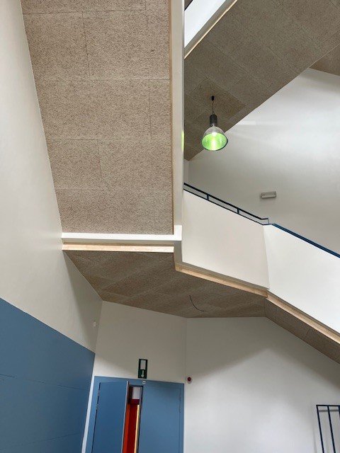 Moderne trap met aan de ene kant textuurrijke wanden van kurk of houtpanelen en aan de andere kant een gladde witte wand, en een groene lamp zichtbaar bovenaan de trap.