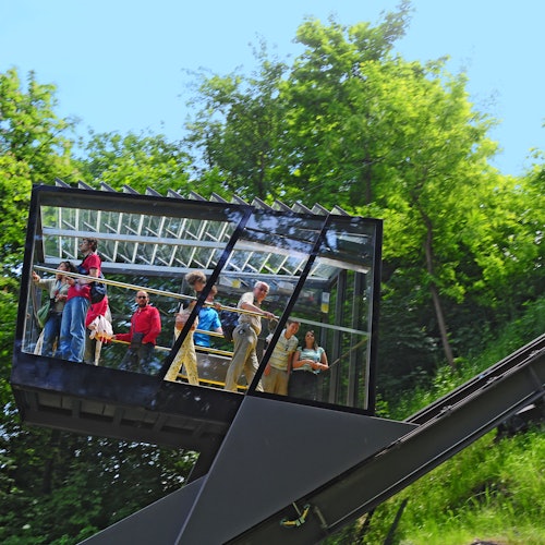People inside a glass-walled funicular car ascending a hillside, with green trees and blue sky in the background.