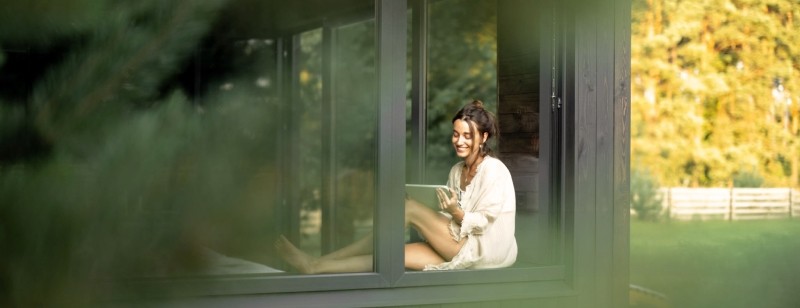 Een jomge vrouw zit in het raamkozijn van een modern huis in een bosrijke omgeving. Haar hele huis is geoptimaliseerd met Huiskracht