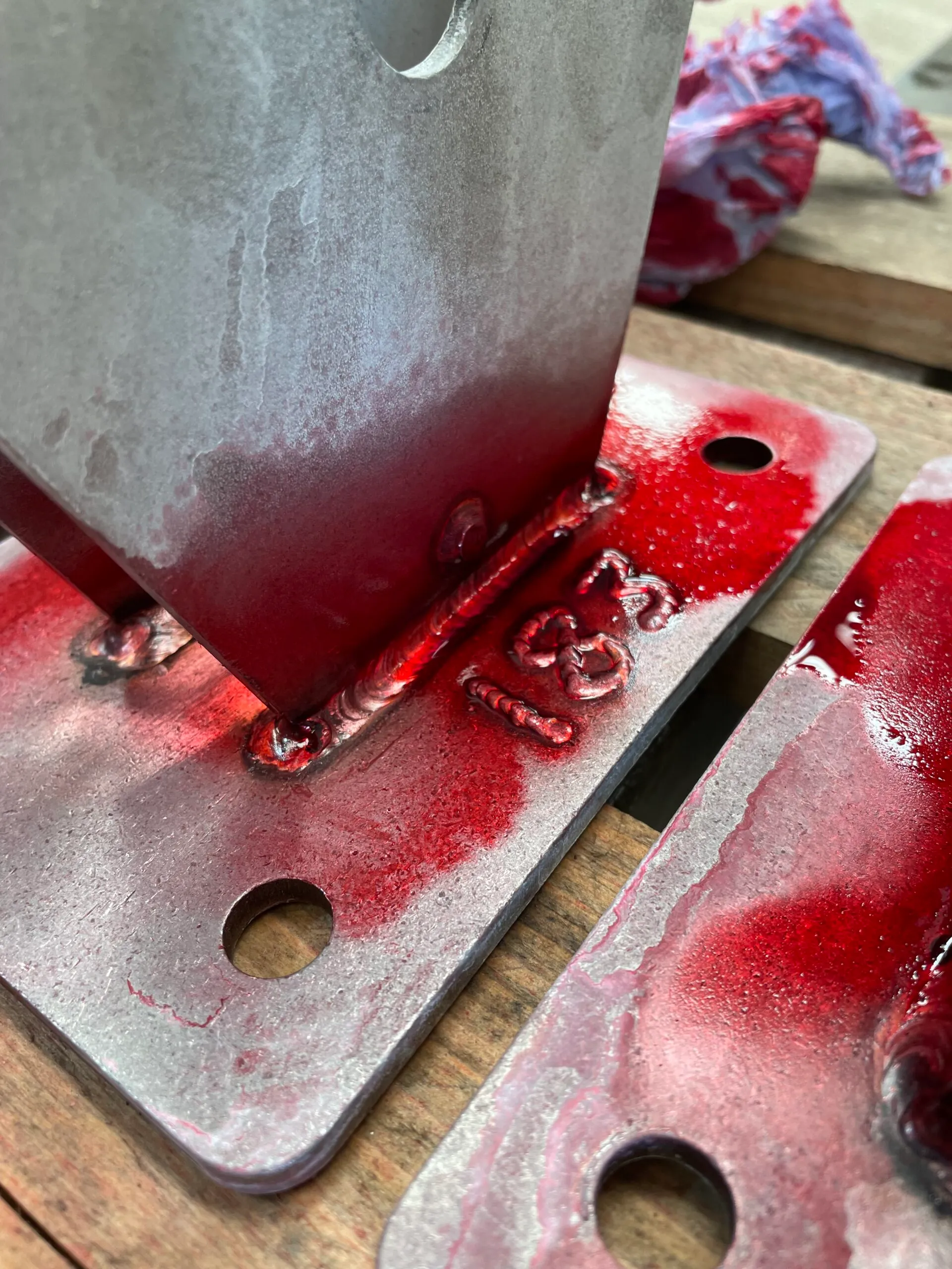 Close-up of metal plates with visible welding seams, coated with red marking dye. The setup is on a wooden surface, exuding an industrial tone.