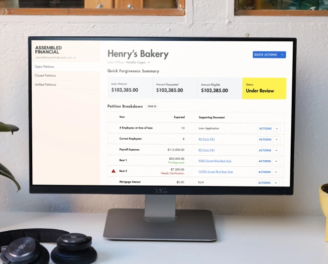 A desktop computer monitor on a desk showing a financial dashboard for 'Henry's Bakery' with a 'Quick Forgiveness Summary' and various financial details.
