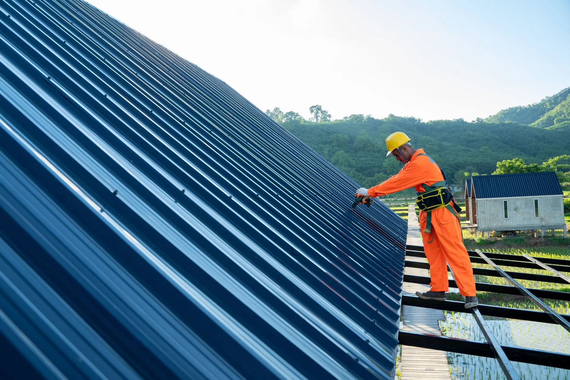 A person wearing gloves uses a yellow pneumatic nail gun to attach dark gray asphalt shingles to a sloped roof.