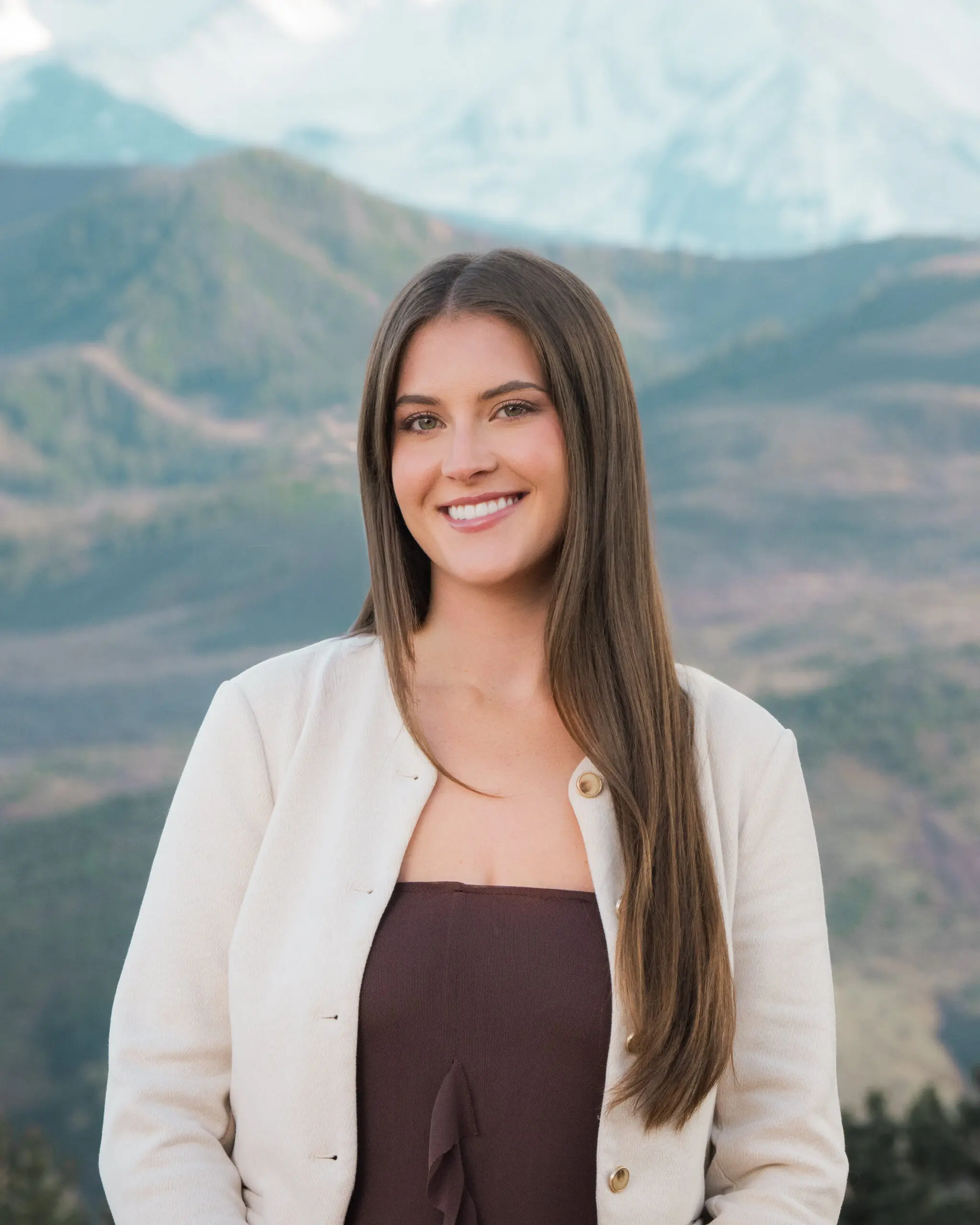 Woman in an ivory jacket and brown top, with mountains behind her.