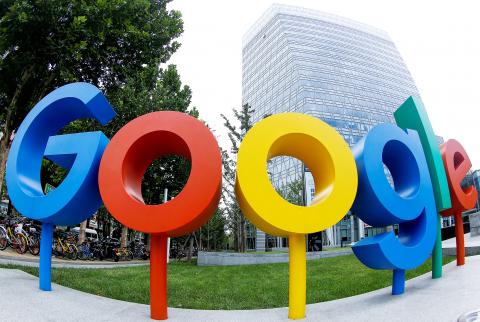 Colorful Google logo sculpture outside a modern glass office building, surrounded by greenery and parked bicycles, symbolizing innovation.