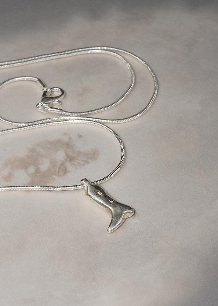 A silver snake chain necklace with a small silver cowboy boot pendant, displayed on a neutral, textured light gray surface.