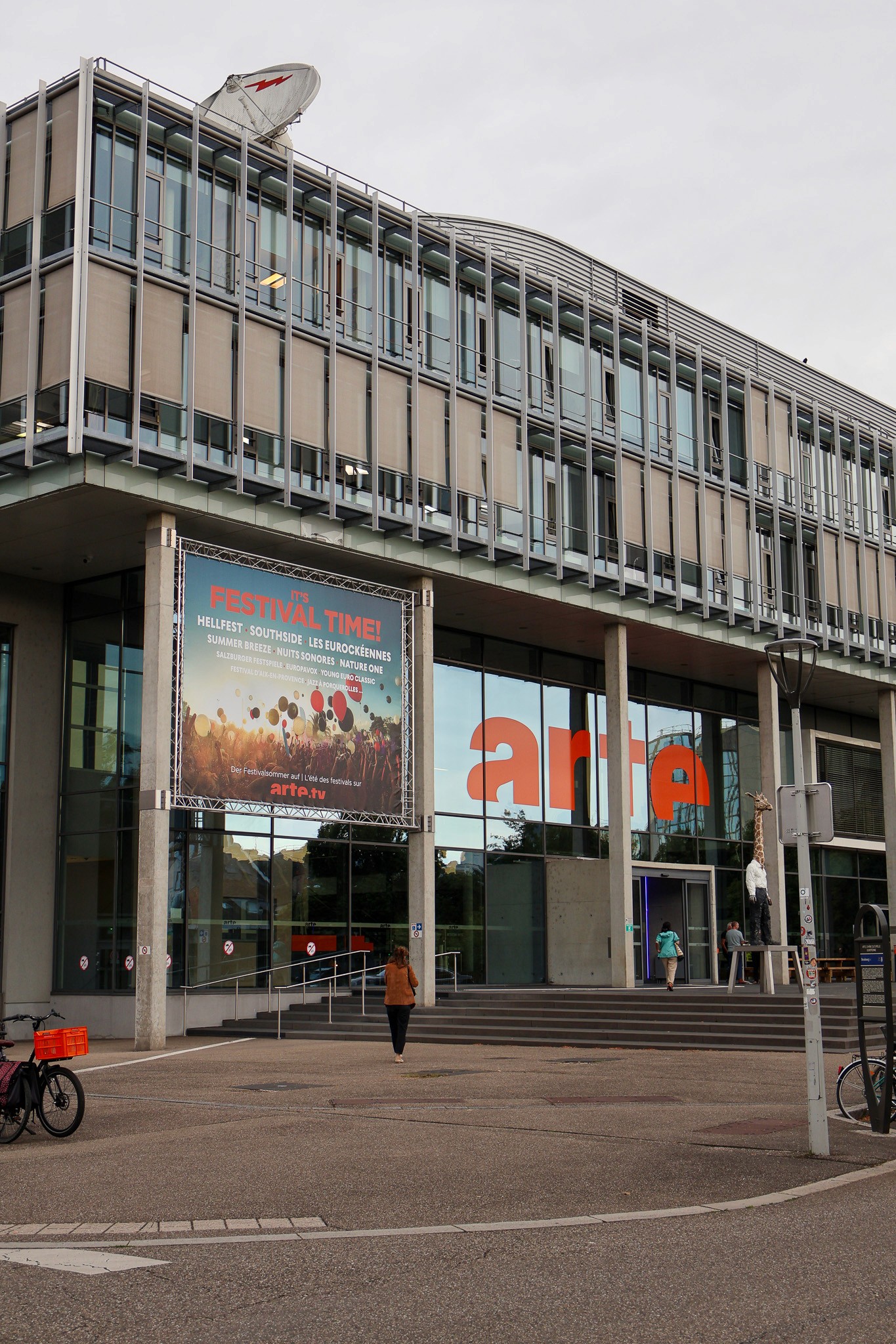 A large banner with the word “Festival Time” written on it, in front of a glass façade of a modern building with the word “arte” on its glass facade and a place in front of it.