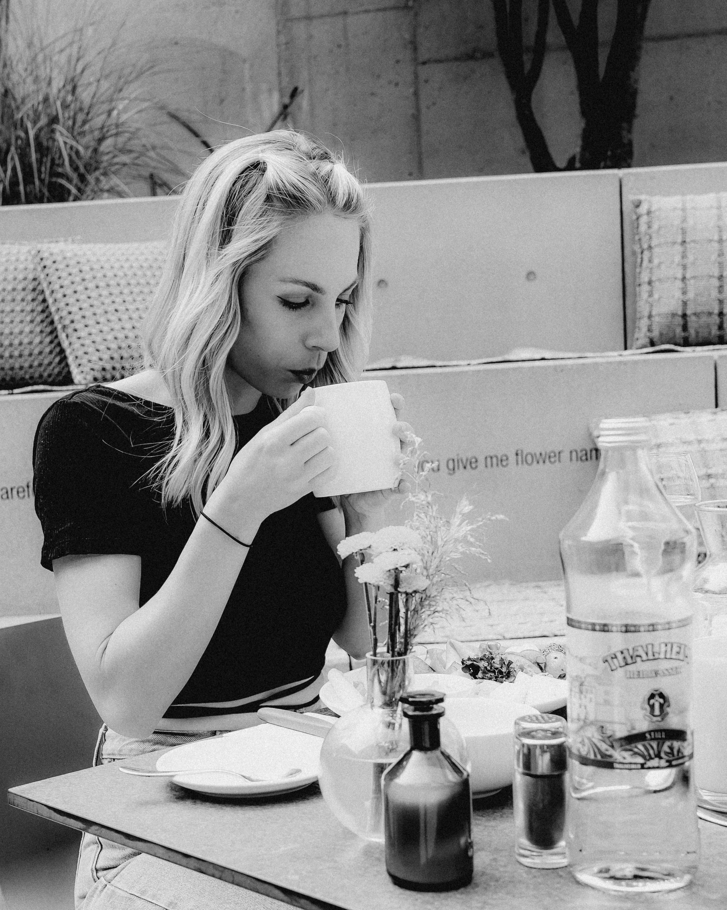 A woman sitting at a table sipping from a mug, with breakfast items in front of her, captured in black and white.