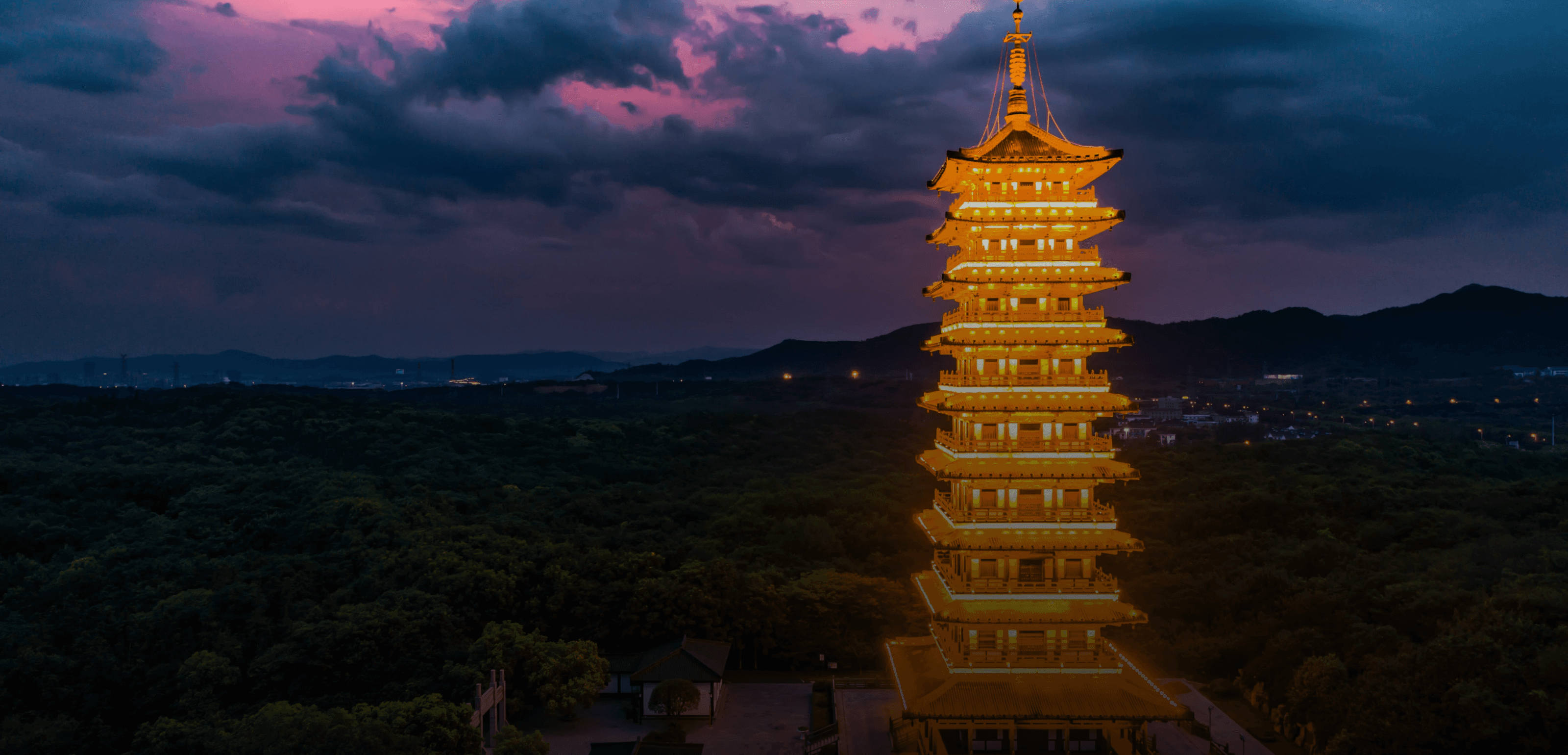 A golden pagoda stands against a dramatic sunset sky, surrounded by lush green hills.
