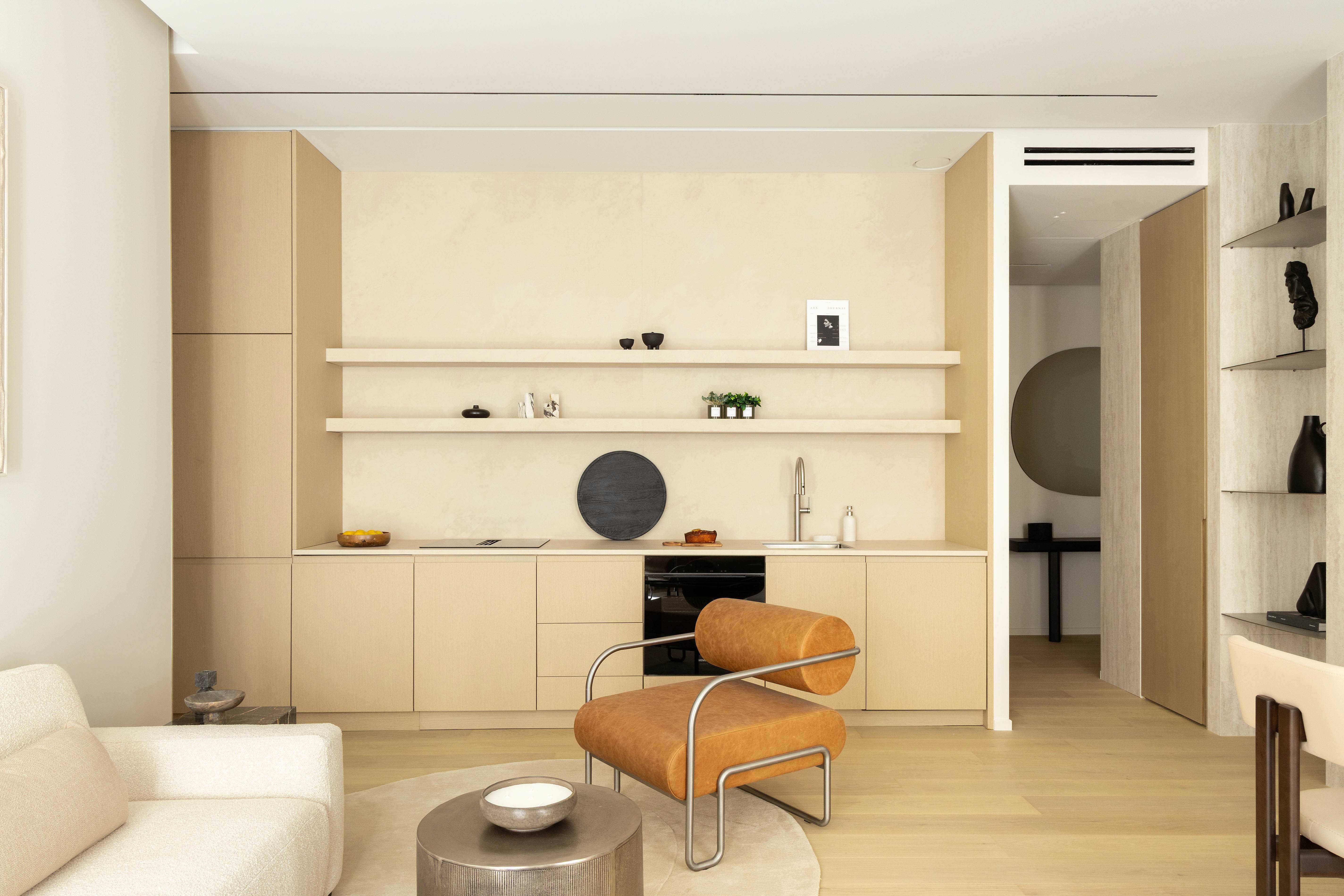 Vue d'ensemble cuisine ouverte avec fauteuil cognac, etageres, four encastre et acces couloir chambres