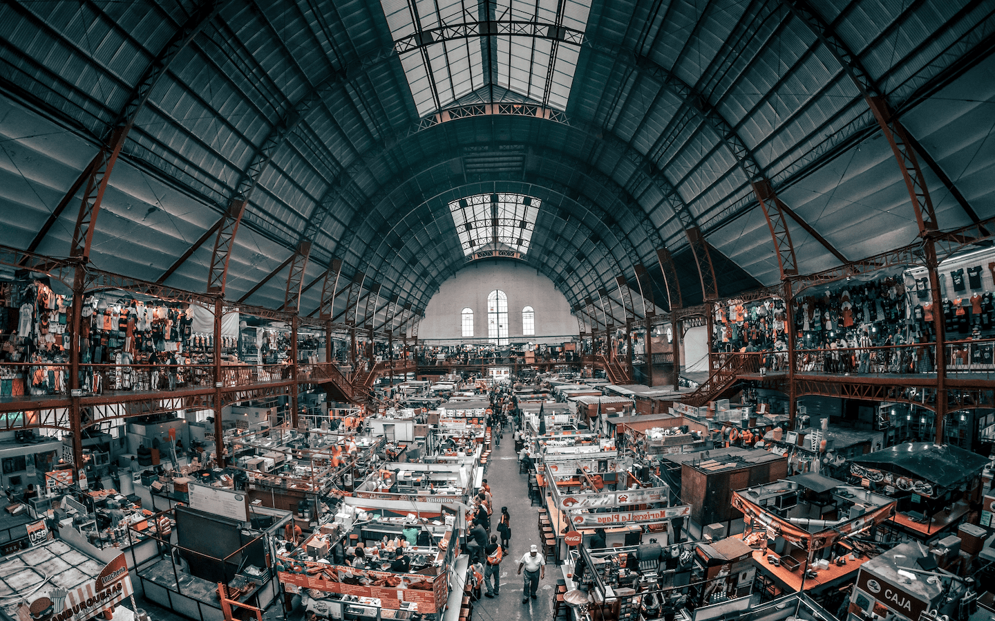 Une animation d'un marché couvert avec de nombreux stands sous un grand toit voûté, où une foule de personnes fait ses achats et se promène.