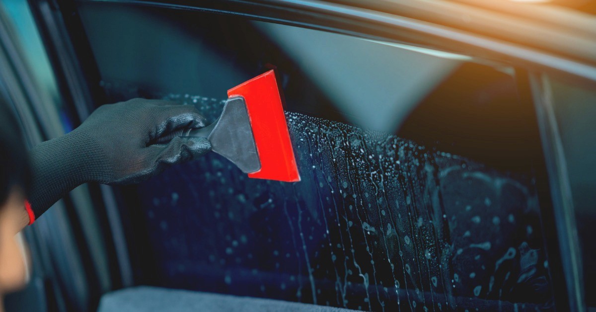 A close-up of a black-gloved hand using a red squeegee to install tinted window film on the side window of a car.