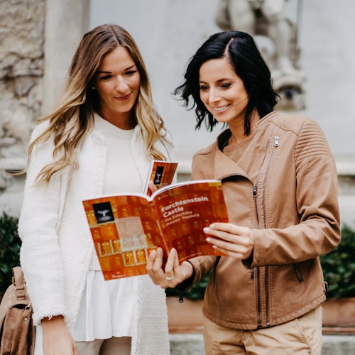 两名女士在户外阅读一本名为《Forchtenstein Castle》的手册。其中一位穿着白色衣服，另一位穿着棕色外套。