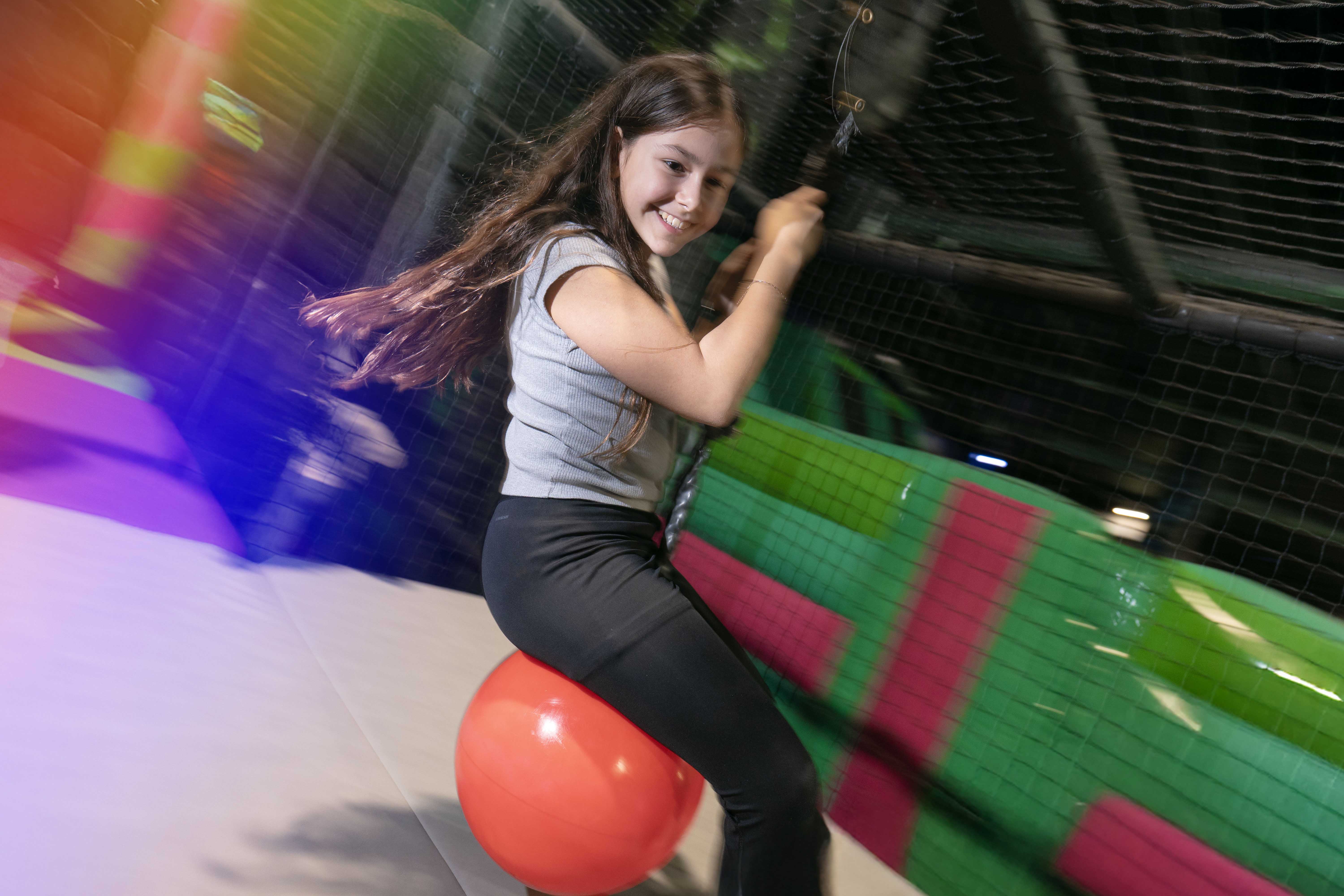 young girl hanging onto swinging ball, play area having fun at Flip Out Adventure Park