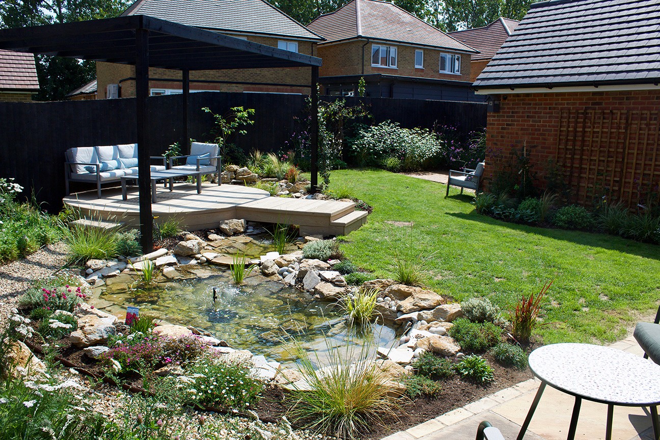 A serene garden with a small pond, rocks, and plants, surrounded by wooden structures and green grass.