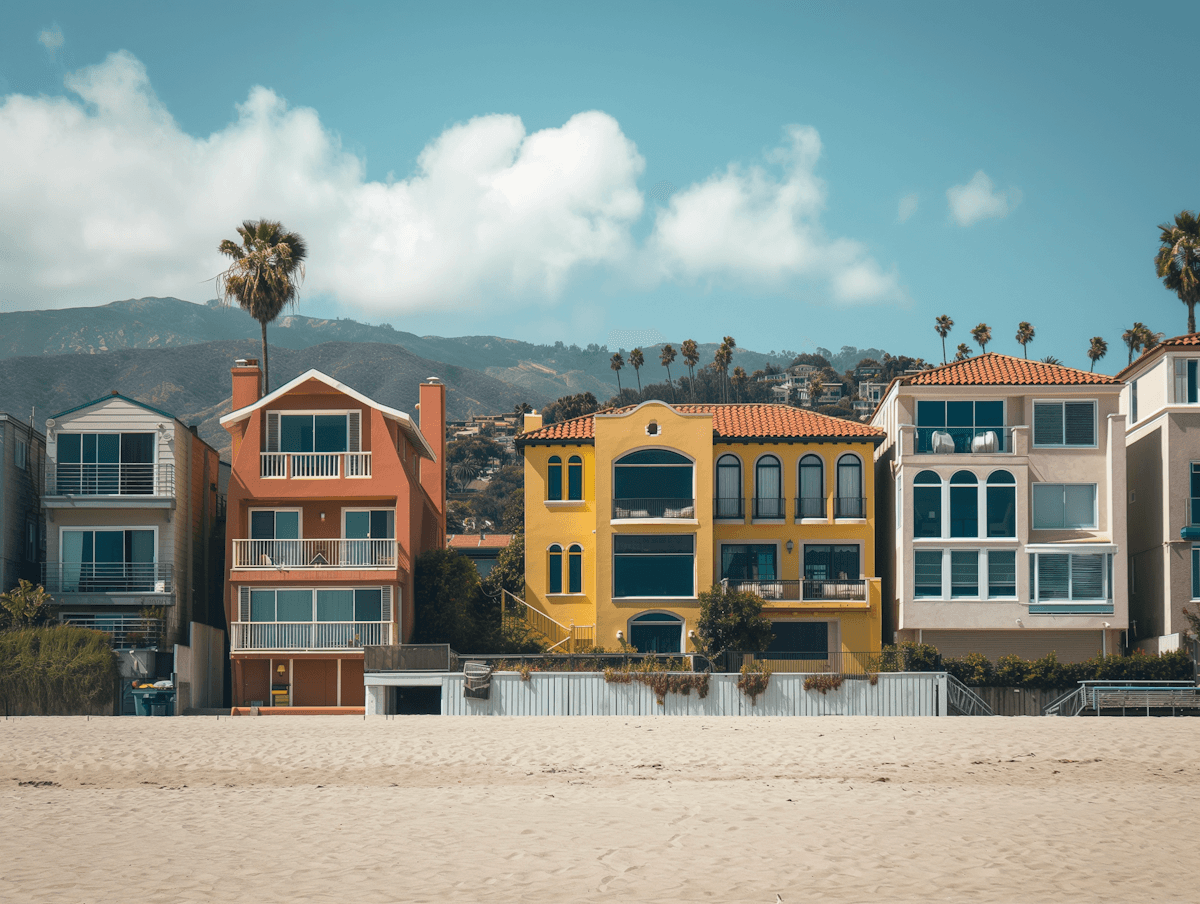 Colorful beach houses with balconies line the sandy shore, backed by mountains and palm trees under a partly cloudy blue sky, evoking a relaxed vibe.