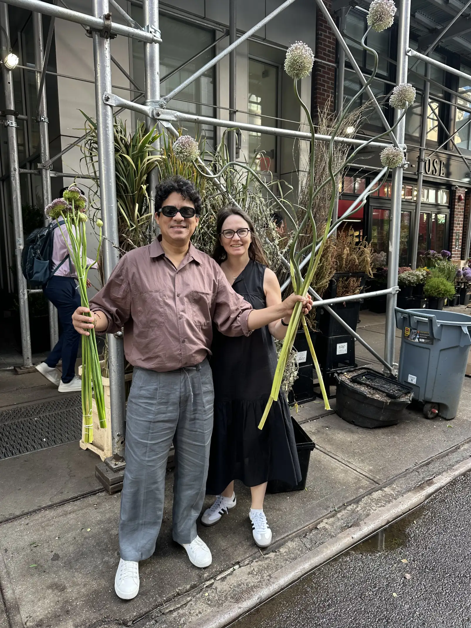 Irina Khatsernova sonrientes posan en las calles de Nueva York sosteniendo tallos largos con flores esféricas, rodeados de estructuras metálicas y puestos de plantas.