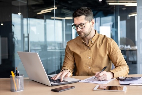 A man with glasses wearing a mustard jacket working at a laptop at an office desk with a glass wall in the background