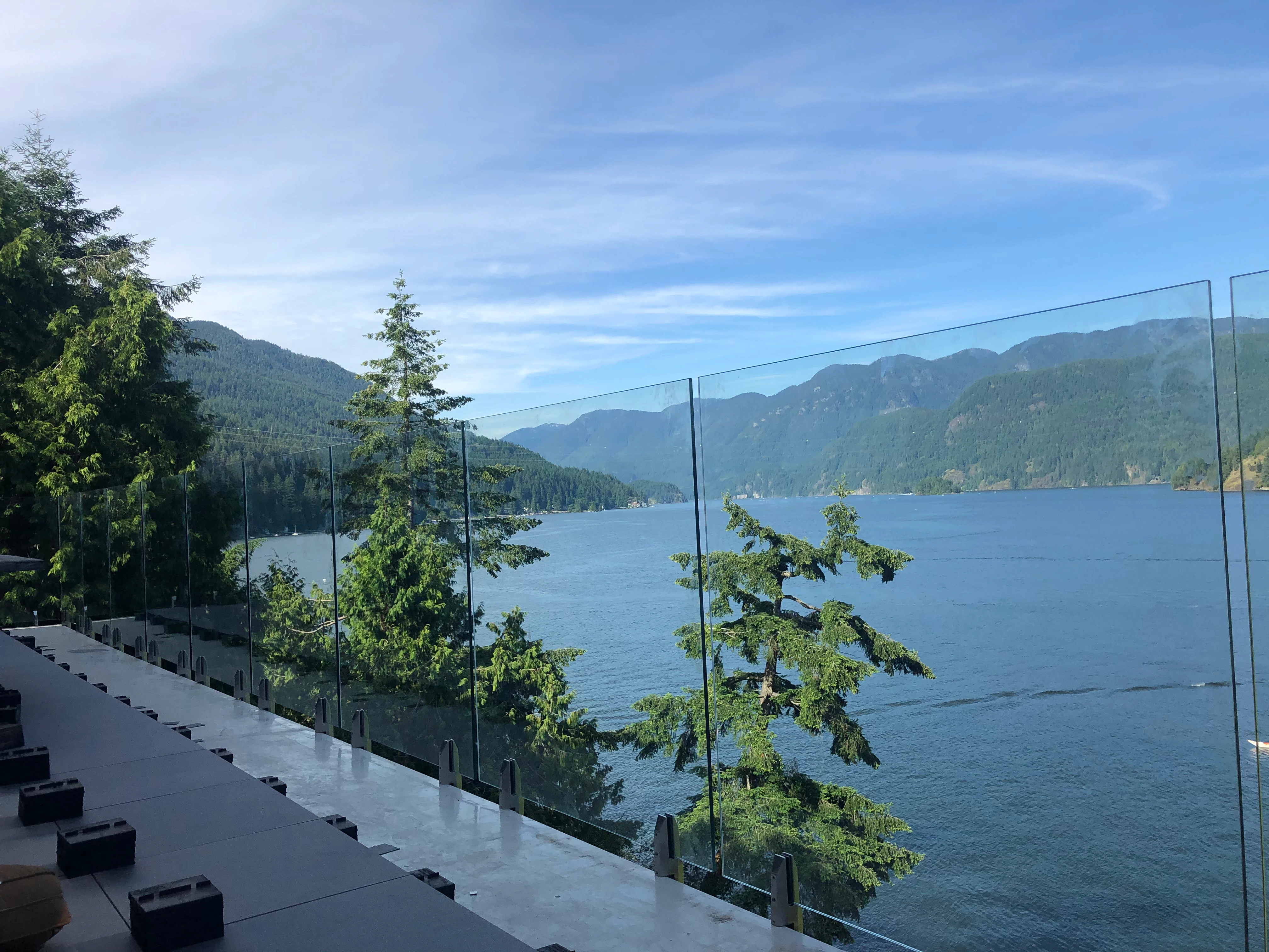 Frameless glass balcony railing overlooking a mountain lake with forested shoreline and panoramic waterfront views under a clear blue sky.