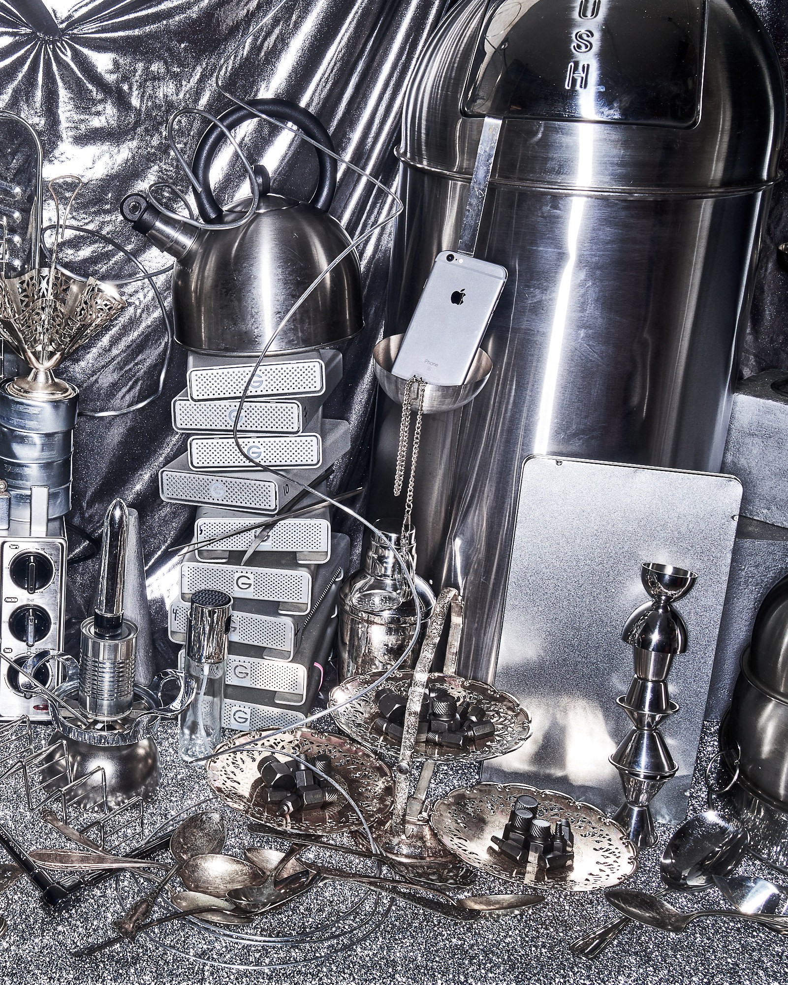 Vertical silver still life assembled from metal vessels, storage drives, electronics, and tableware