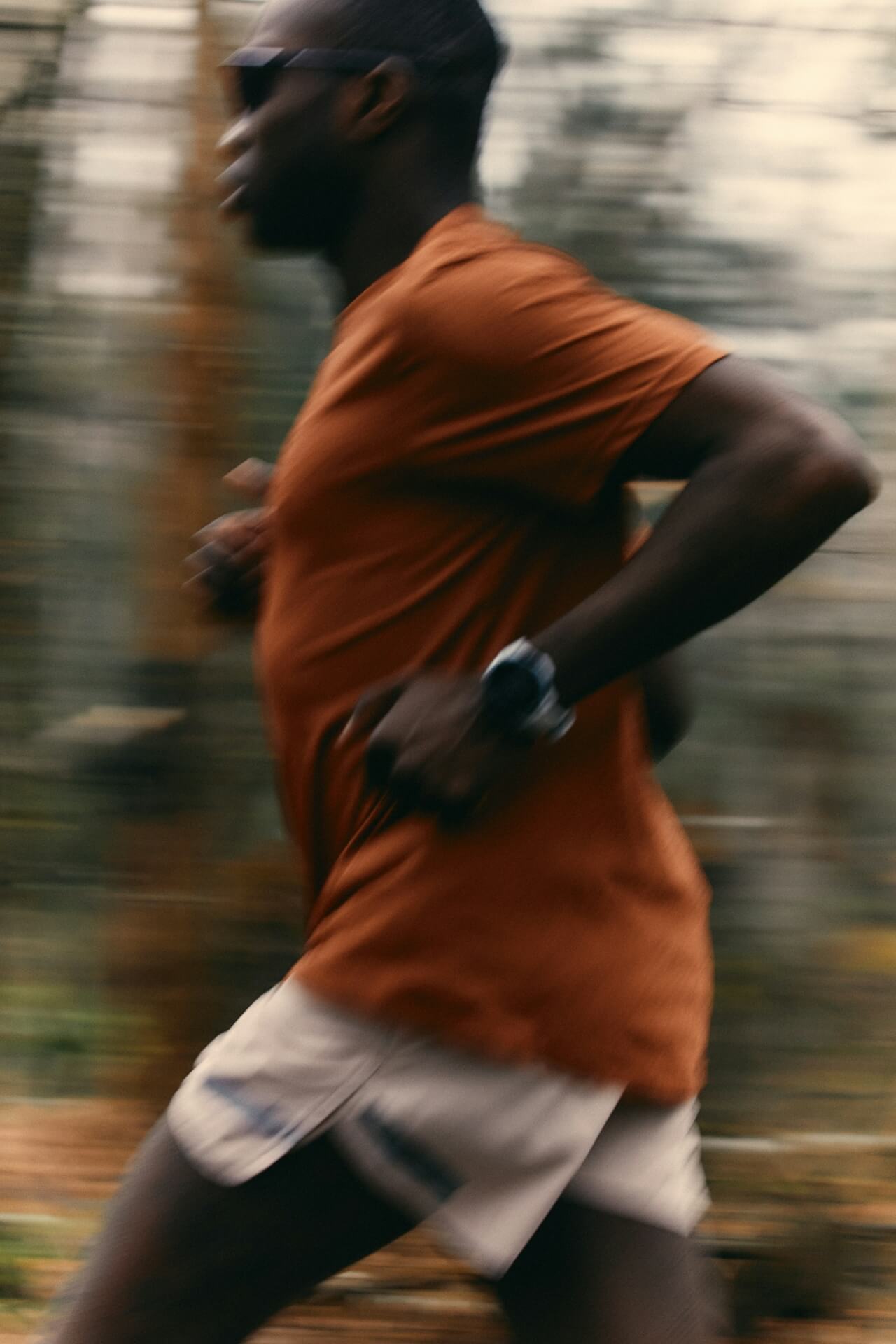 Blurred motion image of a person running in a forest, wearing a brown shirt and shorts. The dynamic scene conveys energy and speed.
