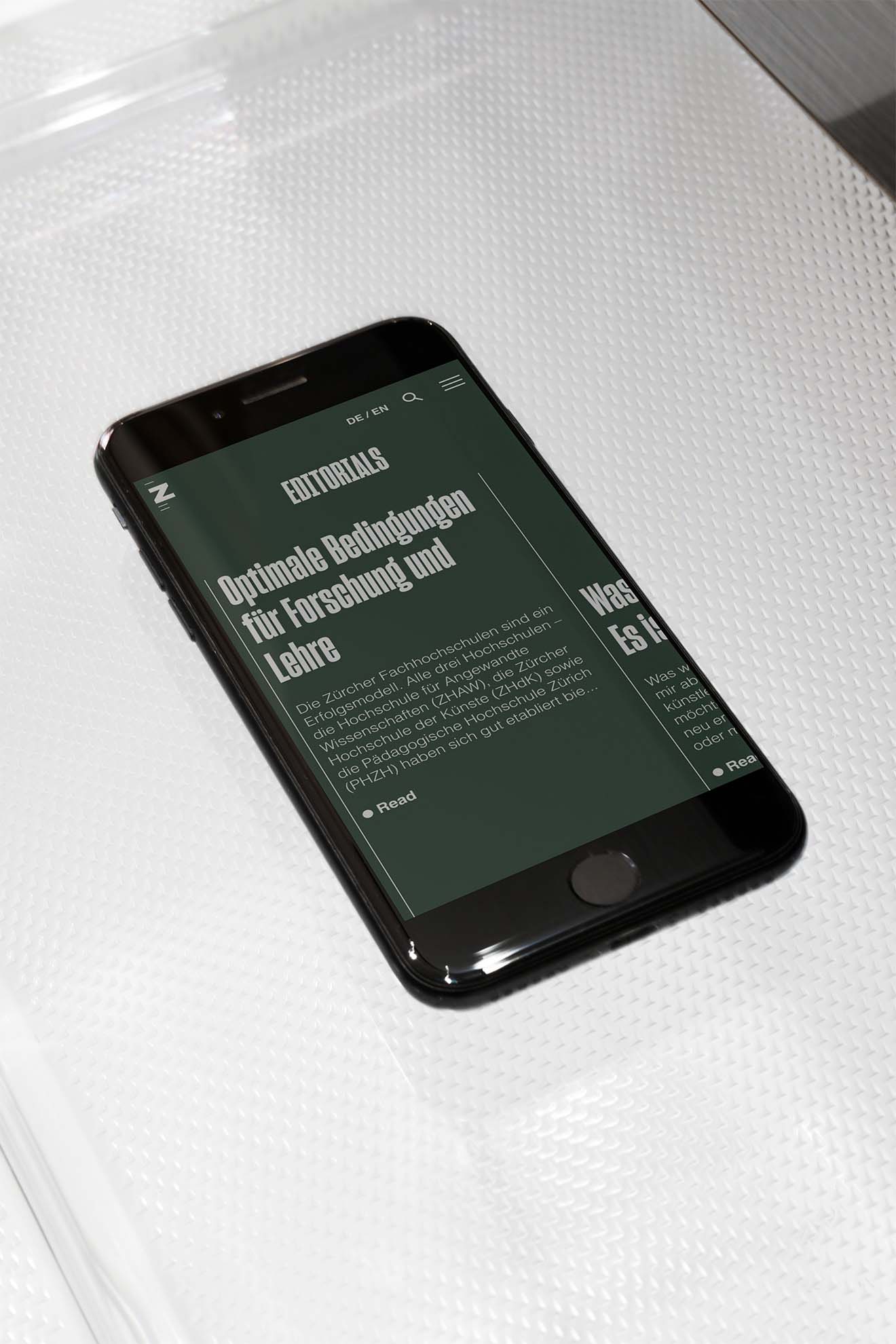 A smartphone resting on a textured white surface shows an editorial page of the ZHdK digital platform. The dark mode UI design features high-contrast white and lime-green text, highlighting a section on "Optimal Conditions for Research and Teaching." This mockup demonstrates a sophisticated content strategy and accessible digital interface created by the Zurich-based agency.